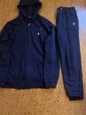 Mörkblå loungewear-set från Polo Ralph Lauren - Mörkblått loungewear-set från Polo Ralph Lauren med dragkedja på hoodien och matchande byxor med dragsko i midjan. Båda delarna har den klassiska loggan broderad i vitt. Helt oanvänd 