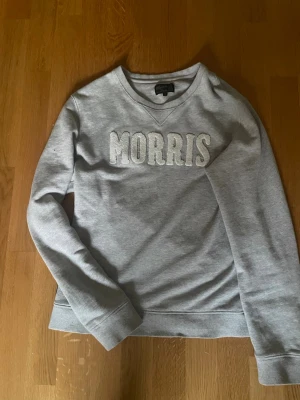 Grå sweatshirt från Morris - Snygg grå sweatshirt från Morris med stor logga broderad på bröstet. Tröjan har rund halsringning, ribbade muddar vid ärmslut och nederkant samt en klassisk passform. Perfekt för en avslappnad och stilren look.