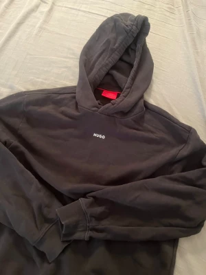 Svart hoodie från Hugo med logga - Snygg svart hoodie från Hugo med liten vit logga på bröstet och klassisk huva med dragsko. Tillverkad i mjukt bomullsmaterial och har ribbade muddar vid ärmslut och nederkant. Storlek S och både kvinnor och män kan använda det och säljer den för bra pris eftersom jag it använder den mer. Pris kan diskuteras 