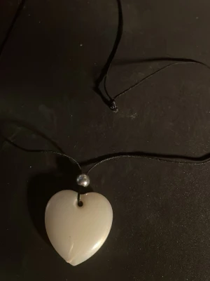 Halsband med hjärthänge och pärldetalj - Snyggt halsband med svart snöre, ett vitt hjärthänge i slät finish och en liten pärlliknande detalj ovanför hänget. Perfekt accessoar för att lyfta din outfit med en enkel men stilren touch.