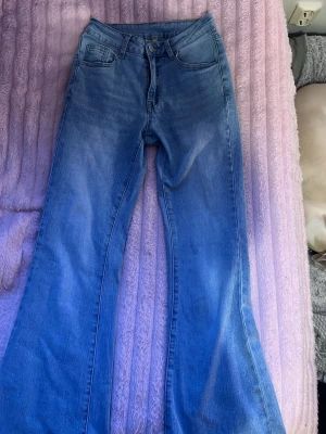 Blå bootcut jeans med hög midja - Säljer ett par ljusblå bootcut jeans med hög midja och klassiska fem fickor. Jeansen har en lätt tvättad look och vida ben som ger en retrovibe. Perfekta för dig som gillar en avslappnad men trendig stil. Materialet är mjukt denim i bomull.