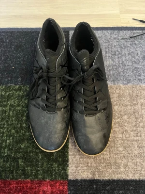 Svarta Adidas fotbollsskor inomhus - Säljer ett par svarta Adidas fotbollsskor för inomhusbruk med klassisk snörning och låg profil. Skorna har en gummisula i ljusbrunt och diskreta mönster på ovandelen. Perfekta för futsal eller inomhusträning.
