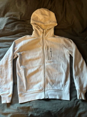 Vit Sail Racing hoodie med dragkedja - Vit hoodie från Sail Racing med hel dragkedja och huva. Modellen har en bröstficka med dragkedja och logotyp på bröstet. Materialet är mjukt och fleeceliknande på insidan, vilket gör den extra bekväm. Perfekt för dig som gillar stilrena och sportiga plagg.