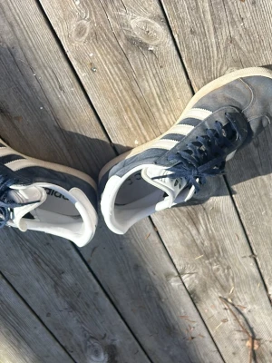 Adidas Gazelle mörkblå sneakers - Klassiska Adidas Gazelle sneakers i mörkblå mocka med vita detaljer och de ikoniska tre ränderna på sidan. Skon har snörning, låg profil och vit sula. Perfekt för dig som gillar retrostil och streetwear.