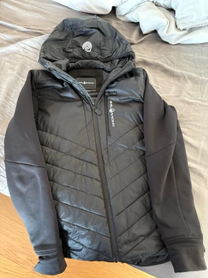 Sail racing cardigen  - Snygg svart vindjacka från sail racing med huva och dragkedja framtill. Jackan har quiltade partier på framsidan och släta ärmar. Perfekt för blåsiga dagar och har en sportig look med logga på bröstet.