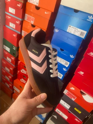 Svarta Hummel sneakers med rosa detaljer - Snygga svarta sneakers från Hummel med klassiska rosa chevrons på sidan och vita skosnören. Modellen heter VM78 CPH och har en brun gummisula som ger retrovibbar. Ovandelen är i mocka och textil, perfekt för dig som gillar streetwear och sportig stil.