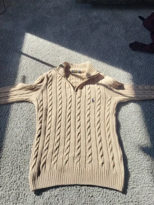 Beige kabelstickad tröja Polo Ralph Lauren - Kabelstickad tröja i beige från Polo Ralph Lauren med klassisk krage och liten broderad logga på bröstet. Tröjan har långa ärmar, ribbade muddar och är gjord i mjukt material som passar perfekt till höst och vinter.