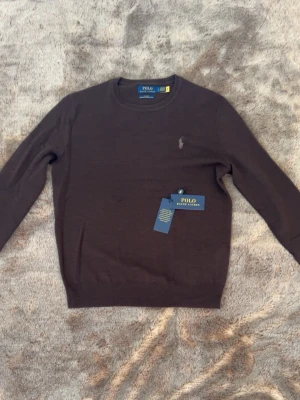  merinoullströja från Polo Ralph Lauren - Mörkbrun stickad tröja från Polo Ralph Lauren i slim fit-modell, tillverkad av tvättbar merinoull. Klassisk rund halsringning och diskret broderad logga på bröstet. Perfekt för dig som gillar stilrena och tidlösa plagg.
