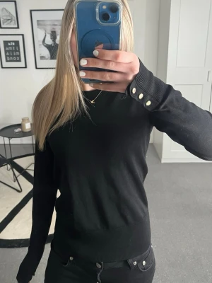 Svart Lindex tröja - Säljer en svart tröja med en kroppad passform från Lindex. Den har tre guldiga knappar på varje arm. Den är som ny men har ett hål längst ner på båda händer (se bild).