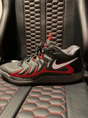 Nike KD LATE NIGHT  - Snygga basketskor från Nike i svart och rött med vit swoosh på sidan. Skorna har lågt skaft, svarta skosnören och en grå sula. Ovandelen är i mesh och syntetmaterial med röda detaljer som ger en sportig vibe.