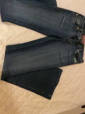 Blå raka jeans från Wrangler - Säljer ett par klassiska blå jeans från Wrangler med raka ben och femficksmodell. Jeansen har en mörk tvätt och detaljerade sömmar vid fickorna. Perfekta för en avslappnad och tidlös stil.