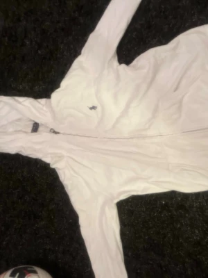 Vit hoodie från Polo Ralph Lauren - Säljer en vit hoodie från Polo Ralph Lauren med dragkedja framtill och klassisk huva. Tröjan har det ikoniska Polo-märket broderat på bröstet och är tillverkad i mjukt material som känns skönt mot huden. Perfekt för en avslappnad och stilren look.