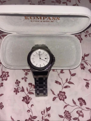 Silverfärgad klocka från Kompass - Stilren klocka från Kompass Stockholm med silverfärgat armband i metall och vit urtavla. Klockan har minimalistisk design med tydliga markeringar och datumvisning. Perfekt accessoar för dig som gillar klassisk och clean stil.