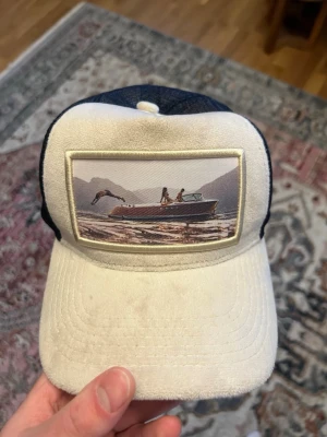 Vit och marinblå keps med bild - Säljer en vit och marinblå keps med ett stort tygmärke framtill som visar en båt på vattnet med berg i bakgrunden. Kepsen har böjd skärm och mesh baktill för extra ventilation. Perfekt för dig som gillar chill stil och naturmotiv.