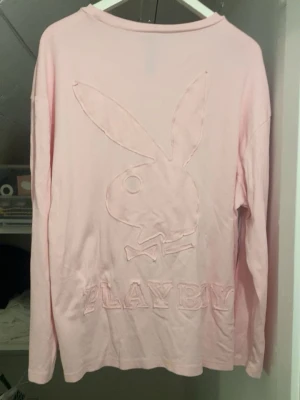 Playboy tröja  - Säljer en ljusrosa långärmad tröja från Playboy med det klassiska kaninlogotypen och texten 'PLAYBOY' i upphöjt tyg på framsidan. Tröjan har en relaxed oversized passform. Liten fläck (bild 2) 