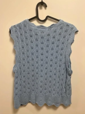 Ljusblå stickad väst med hålmönster - Supersöt ljusblå stickad väst med vågig kant och detaljerat hålmönster över hela plagget. Ärmlös modell med rund halsringning, perfekt att styla över en skjorta eller t-shirt. Materialet känns mjukt och luftigt, vilket gör den skön att bära under varmare dagar.