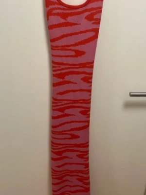 Rosa och röd zebramönstrad långklänning - Cool långklänning i rosa och rött med zebramönster. Klänningen är ärmlös och har en rund halsringning. Materialet ser ut att vara stickat och stretchigt, vilket ger en slim passform. Perfekt för dig som vill sticka ut med färg och mönster.