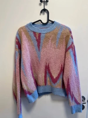 Pastellfärgad stickad tröja - Mysig stickad tröja i pastellfärger med zigzag-mönster i rosa, blått, beige och vinrött. Tröjan har rund halsringning, ribbade muddar och avslut. Perfekt för dig som gillar färg och unik stil.