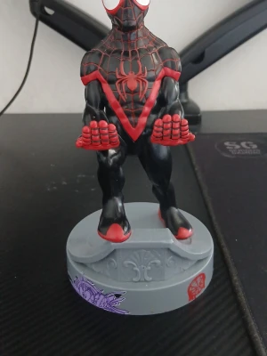 Miles Morales Spiderman figur  En mobil eller kontrollhållare - Cool samlarfigur av Miles Morales Spiderman i svart och rött med tydliga spindelnätsdetaljer och stor röd spindel på bröstet. Figuren står på en rund grå bas med lila och röda mönster. Perfekt för skrivbordet eller hyllan.