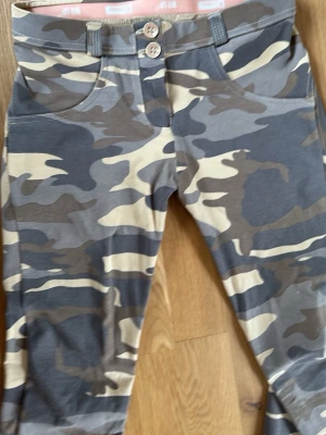 Cargobyxor i camomönster från Freddy WR UP - Snygga cargobyxor från Freddy med klassiskt camomönster i beige, grått och grönt. Byxorna har två knappar framtill, bälteshällor och mjukt stretchigt material för extra komfort. Perfekta för en avslappnad streetwear-look.