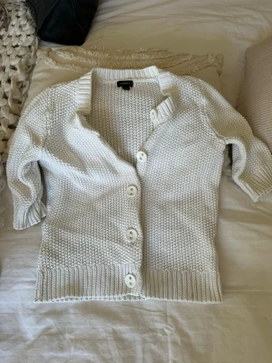 Vit stickad kofta från Lindex - Vit stickad kofta från Lindex med grovstickad struktur, stora vita knappar och ribbade muddar. Koftan har halvlånga ärmar och en klassisk krage. Perfekt att slänga över en topp för en chill look.