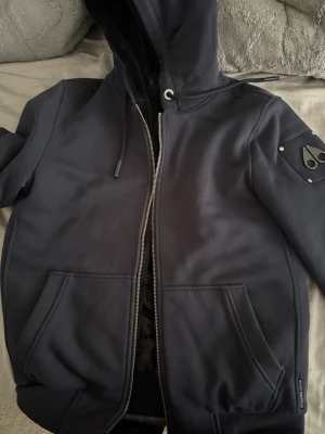 Mörkblå  hoodie från Moose Knuckles - Säljer en svart hoodie från Moose Knuckles med dragkedja, stor huva och två rymliga fickor framtill. Ärmen har den ikoniska metall-loggan på en patch med nitar. Tillverkad i mjukt och tjockt material som känns riktigt lyxigt.ÄKTA säljer den eftersom det är inte min stil längre. Vill sälja denna snabbt då jag behöver pengar . Som ny använt 2-3 gånger inga håll eller nått annat du kommer bli nöjd . Storlek S men passar M .jag har M och den passar perfekt 