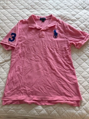 Rosa piké från Ralph Lauren - Säljer en klassisk rosa pikétröja från Ralph Lauren med blå logga på bröstet och siffran 3 på ärmen. Tröjan har krage, korta ärmar och är tillverkad i bomull. Perfekt för en avslappnad men ändå stilren look. Den är använd men inte många gånger. Det står att det är xl men det är lite mindre