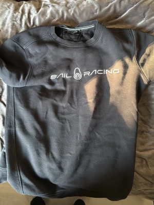 Svart Sail Racing sweatshirt - Svart sweatshirt från Sail Racing med vit broderad logga på bröstet. Klassisk rund halsringning och ribbade muddar. Perfekt för en avslappnad och sportig stil.