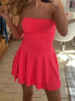 Axelbandslös rosa kortklänning - Snygg axelbandslös kortklänning i stark rosa färg. Klänningen har en figurnära överdel och ett utställt kjolparti som ger en lekfull look. Perfekt för sommarens alla tillfällen. Materialet känns mjukt och stretchigt.