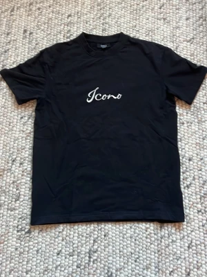 Svart Icono t-shirt - Snygg svart t-shirt från New Yorker med vit text 'Icono' fram och 'Icono Couture' bak. Baksidan har även ett stort röd-svart mönster som ger en unik look. Klassisk passform och korta ärmar, perfekt till jeans eller shorts.