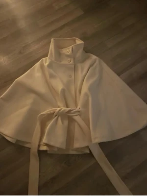 Beige Gina tricot poncho - Stilren beige cape med hög krage och knappar framtill. Knytband i midjan ger en snygg siluett. Capen har en vid, svepande form och är tillverkad i ett mjukt tyg som känns lyxigt. Perfekt för dig som vill ha en unik och trendig ytterrock. Bilden är lånad men skriv för fler