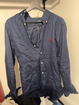 Blå linneskjorta från Polo Ralph Lauren - Säljer en mörkblå skjorta i linne från Polo Ralph Lauren med klassisk button-down krage och röd broderad logga på bröstet. Skjortan har långa ärmar och vita knappar framtill. Perfekt för en avslappnad men ändå stilren look. Storlek S men passar upp mot M. Modellen är 182cm 73 kg