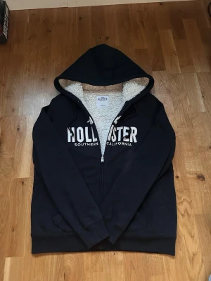 Marinblå hoodie från Hollister - Mysig marinblå hoodie från Hollister med dragkedja och stor vit logga på bröstet. Insidan är fodrad med mjuk teddy för extra komfort. Klassisk huva med dragsko och två fickor framtill. Perfekt för dig som gillar avslappnad och stilren streetwear. L sitter mer som M
