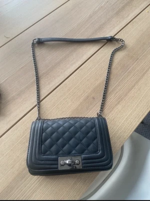 Svart quiltad axelväska med kedja - Snygg svart axelväska med quiltat mönster och silverfärgad kedjerem. Väskan har en rektangulär form och ett metallspänne framtill. Perfekt för dig som gillar stilrena accessoarer med en lyxig touch. Materialet är konstläder.