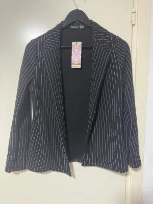 Svart kavaj med vita ränder Boohoo - Snygg svart kavaj från Boohoo med tunna vita vertikala ränder. Klassisk krage och figurnära passform. Perfekt för dig som gillar stilrena och trendiga plagg. Tillverkad i ett mjukt material som känns bekvämt hela dagen.