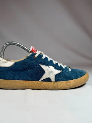 Golden Goose Superstar - Snygga blå sneakers från Golden Goose med vit stjärndetalj på sidan, vita skosnören och beige sula. Ovandelen är i mocka och har en röd GGDB-logga på plösen. Perfekta för dig som gillar en avslappnad och trendig stil. Passar storlek 44