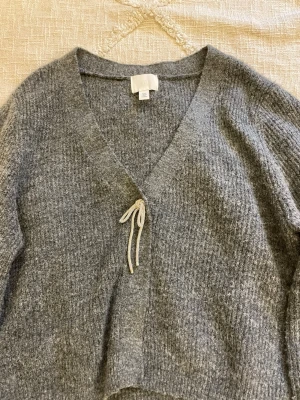 Grå stickad kofta  - Mysig grå stickad kofta från H&M med v-ringning och rosett framtill. Koftan har lång ärm och är gjord i ett mjukt, fluffigt material som känns lyxigt. Säljer pågrund av att tröjan ej kommit till användning💓