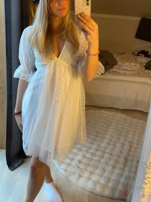 Vit kortklänning med volang och spets - Söt vit kortklänning med v-ringning och volangdetaljer. Klänningen har genomskinliga puffärmar i trekvartslängd och är dekorerad med små spetsprickar. Perfekt för sommarens alla tillfällen.