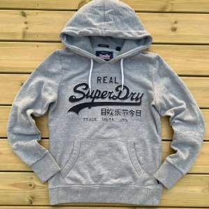 Grå Superdry Hoodie  - Snygg grå hoodie från Superdry med stor svart logga och japansk text på bröstet. Klassisk känguruficka framtill, ribbade muddar och vit dragsko i huvan. Kom privat för mer info och om du har frågor är det bara att ställa!🤙🏽