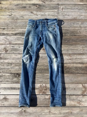 Blå slitna jeans från replay - Säljer ett par blå jeans från Nudie Jeans med skinny fit och slitningar på knäna. Jeansen har klassisk femficksdesign, dragkedjegylf och tydliga slitningar för en cool, använd look. Tillverkade i denim med ljusare tvättade partier. Strl 30-34