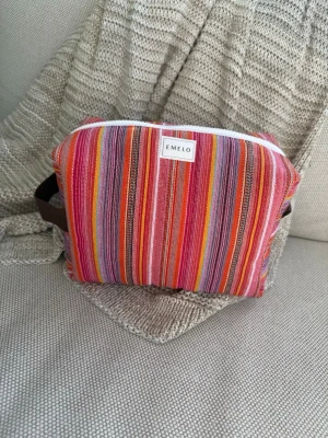 Randig necessär från Emelo - Färgglad necessär från Emelo med ränder i rosa, rött, orange, lila och gult. Väskan har en mjuk, boxig form och är tillverkad i tyg med ett vitt blixtlås och bruna handtag på sidorna. Perfekt för att organisera småprylar eller smink i väskan. Mått ca 26x17x12 cm.