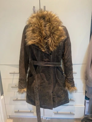 Jofama kappa - Snygg brun kappa i mocka med fuskpälsdetaljer vid krage och ärmar. Kappan har ett matchande bälte i midjan och en figurnära passform. Perfekt för kalla dagar när du vill vara både varm och trendig.