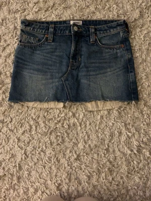 Blå jeanskjol från Monki - Säljer en blå jeanskjol från Monki med rå kant nertill och klassiska fem fickor. Kjolen har en kort passform och är gjord i denim med synliga sömmar. Perfekt för dig som gillar en avslappnad och trendig stil.