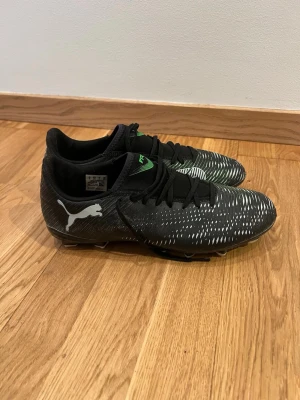 Svarta Puma fotbollsskor med dobbar - Säljer ett par svarta fotbollsskor från Puma med vita och gröna detaljer. Skorna har snörning och slitstark syntetisk ovandel samt fasta vita dobbar för bra grepp på planen. 
