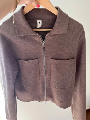 Brun zip cardigan med fickor - Snygg brun cardiganjacka från Åhléns med dragkedja framtill och två stora utanpåfickor. Jackan har en klassisk krage och är tillverkad i ett mjukt, stickat material. Perfekt för lager på lager och enkel att matcha med olika outfits.