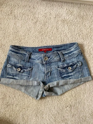 Blå vintage jeansshorts från Gina Tricot - Snygga blå vintage jeansshorts från Gina Tricot med uppvikta benslut och dekorativa fickor med knappar både fram och bak. bälteshällor och knappgylf. Perfekta för varma dagar och enkel att matcha.