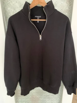 H&M svart half-zip hoodie - En svart hoodie med half-zip dragkedja och hög krage från H&M. Tröjan har en avslappnad passform, ribbade muddar vid ärmslut och nederkant. Tillverkad i mjukt sweatshirtmaterial, perfekt för en chill och stilren look. Oanvänd.