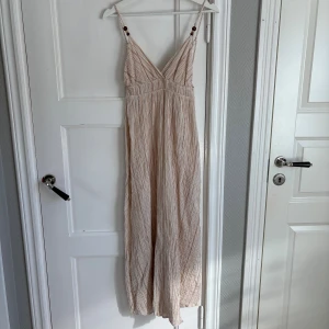 Långrandig omlottklänning med smala band - Långklänning i ljusbeige med tunna bruna ränder och omlottdesign. Klänningen har smala axelband med dekorativa träknappar och v-ringad urringning. Tillverkad i ett luftigt bomullsmaterial, perfekt för varma dagar.