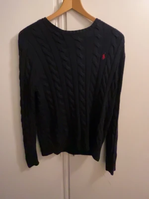 Mörkblå kabelstickad tröja Polo Ralph Lauren - Snygg mörkblå kabelstickad tröja från Polo Ralph Lauren med röd broderad logga på bröstet. Tröjan har rund halsringning och långa ärmar. Perfekt för dig som gillar klassisk och tidlös stil.