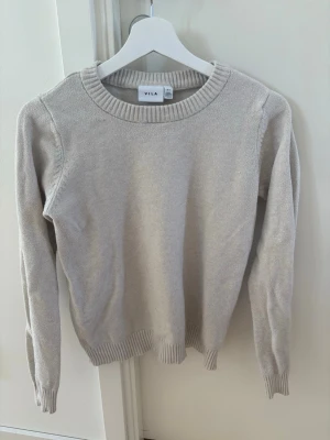 Beige stickad tröja från VILA - Säljer en enkel och stilren beige stickad tröja från VILA. Tröjan har rund halsringning, långa ärmar och ribbade muddar vid ärmslut och nederkant. Perfekt basplagg till garderoben och lätt att matcha med olika outfits.🤍